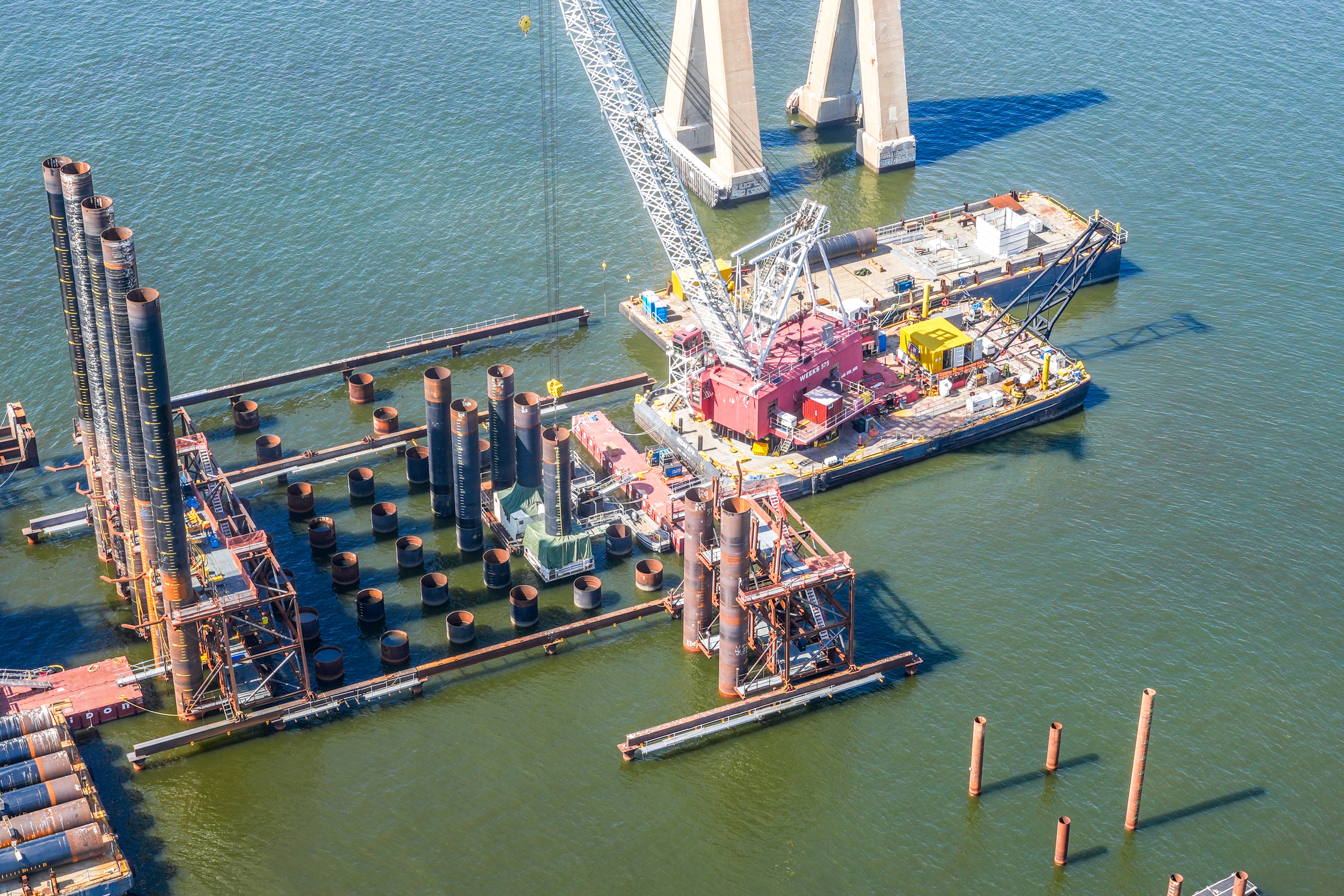 Aerial view of crane on barge installing permanent foundation piles in water.