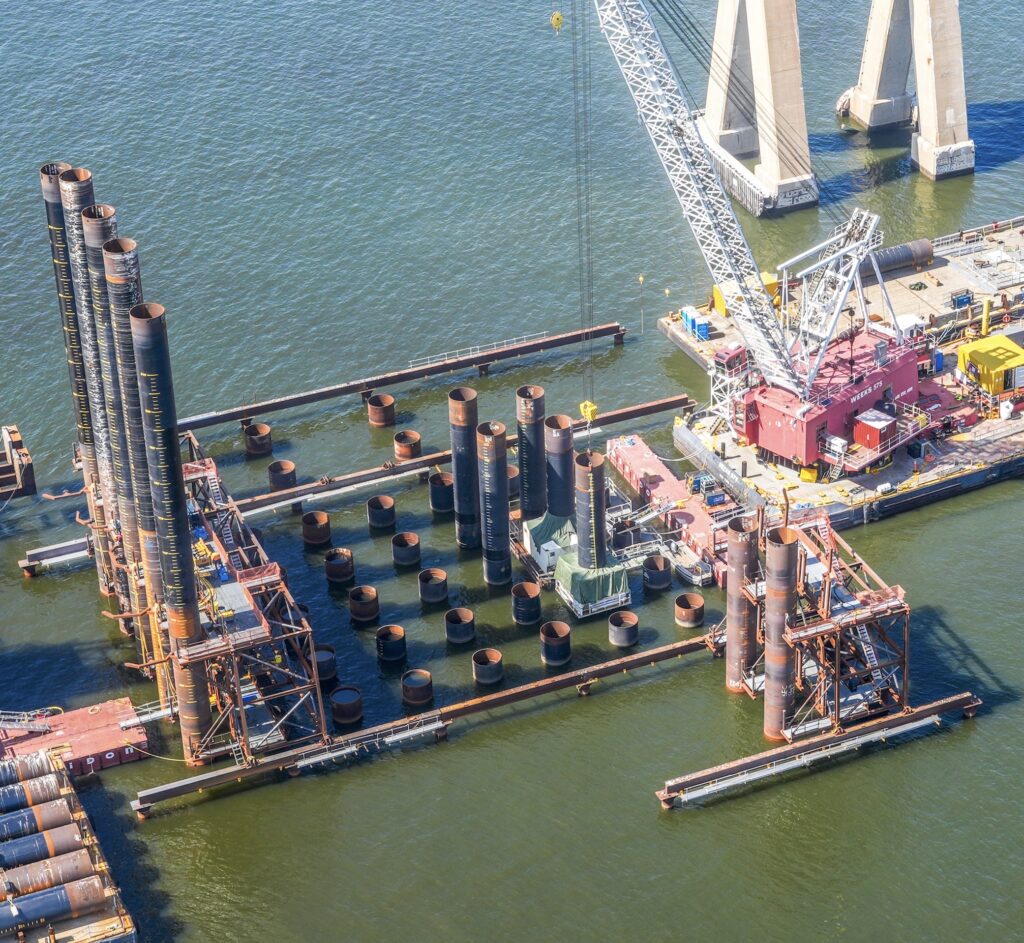 Aerial view of crane on barge installing bridge piles in water.